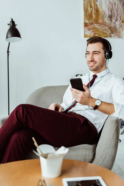 Bel homme d'affaires écoutant de la musique dans les écouteurs et utilisant un smartphone dans le bureau — Photo de stock