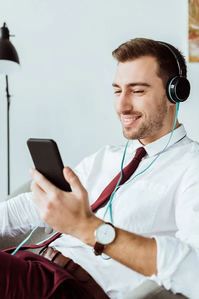 Homme d'affaires souriant écoutant de la musique dans un casque et utilisant un smartphone — Photo de stock