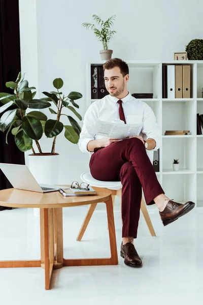 Handsome stylish businessman with newspaper and laptop sitting in modern office — Stock Photo