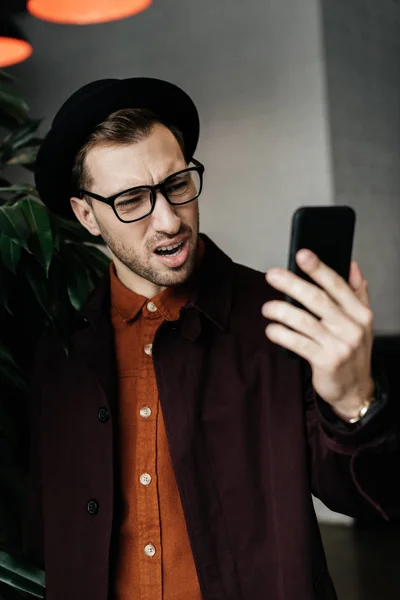 Красивый напряженный мужчина в шляпе и очках, смотрящий на смартфон — стоковое фото