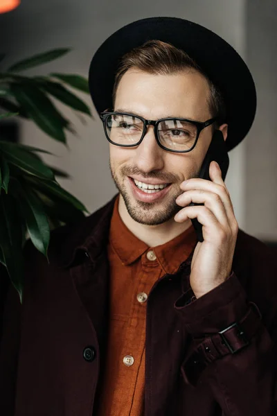 Handsome cheerful man in eyeglasses and hat talking on smartphone — Stock Photo