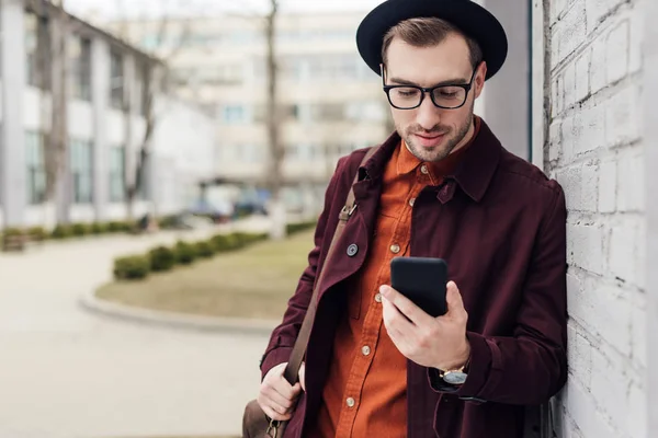 Handsome stylish man in trendy clothes with bag using smartphone — Stock Photo