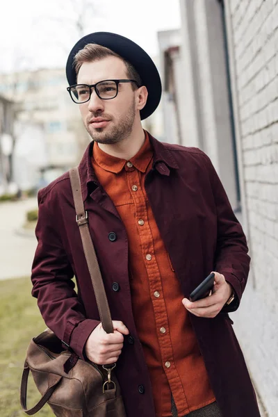 Bel homme élégant dans le chapeau et les lunettes avec sac tenant smartphone — Photo de stock
