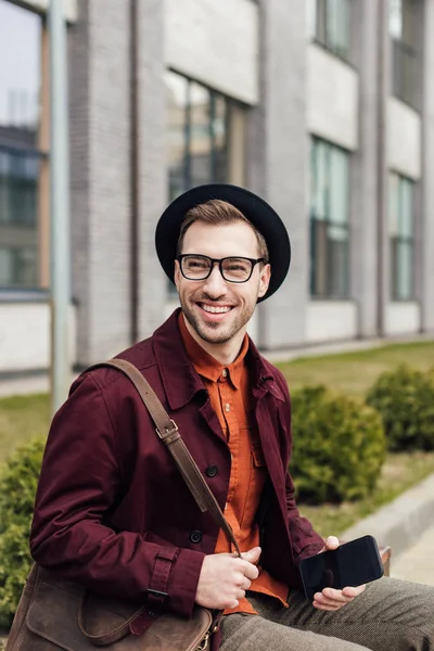 Hombre alegre guapo con bolsa usando teléfono inteligente - foto de stock