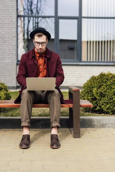 Handsome fashionable man using laptop and sitting on bench — Stock Photo