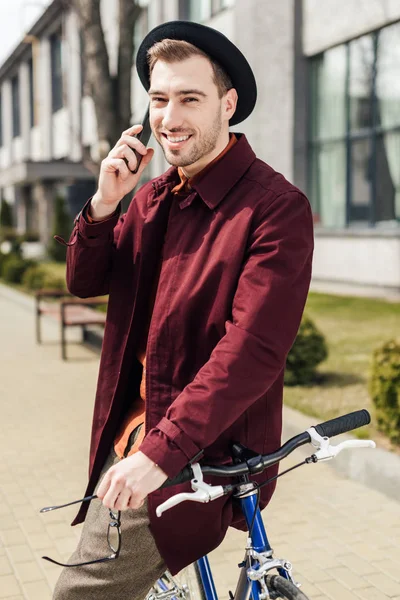 Стильный мужчина в очках и шляпе разговаривает на смартфоне, опираясь на велосипед — стоковое фото