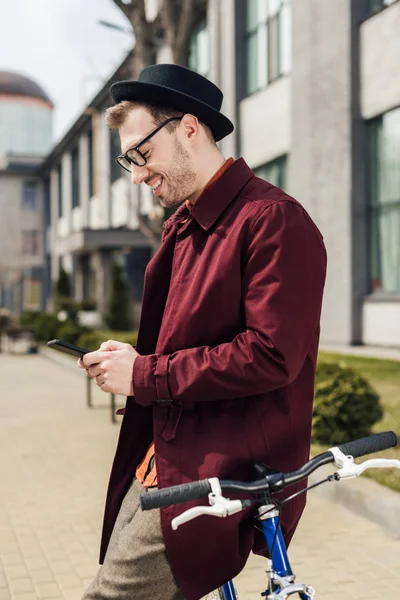 Schöner, stylischer Mann mit Brille und Smartphone, während er sich aufs Fahrrad lehnt — Stockfoto