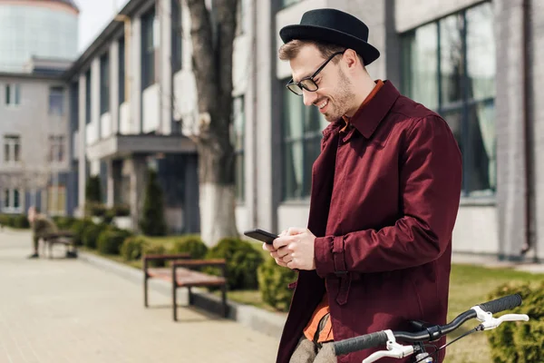 Happy handsome man in eyeglasses using smartphone while leaning on bicycle — Stock Photo