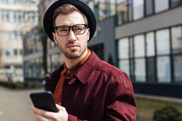 Bel homme sérieux dans les lunettes et le chapeau en utilisant un smartphone — Photo de stock