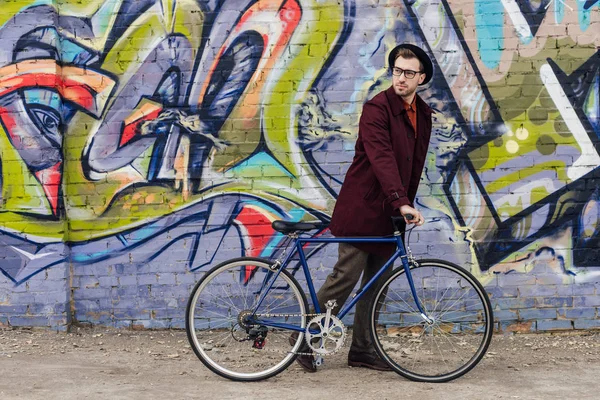 Élégant homme élégant debout avec vélo au mur avec graffiti — Photo de stock