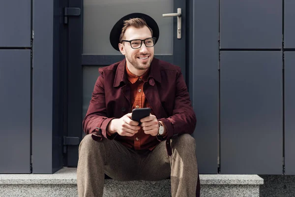 Smiling man in eyeglasses and hat using cellphone — Stock Photo