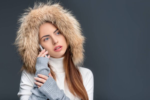 beautiful woman in fur hat and winter outfit talking on smartphone, isolated on grey