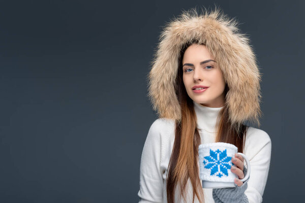beautiful woman in fur hat and winter outfit holding cup of coffee, isolated on grey