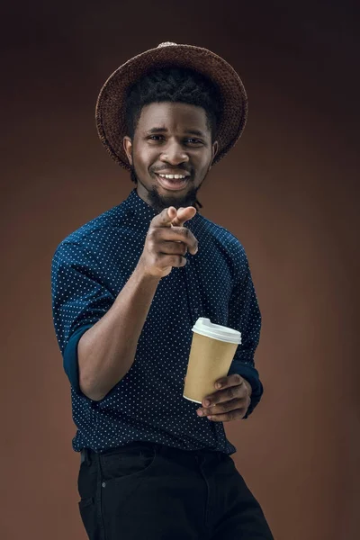 Uomo africano americano con tazza di caffè usa e getta che punta sulla macchina fotografica isolata su marrone — Foto Stock