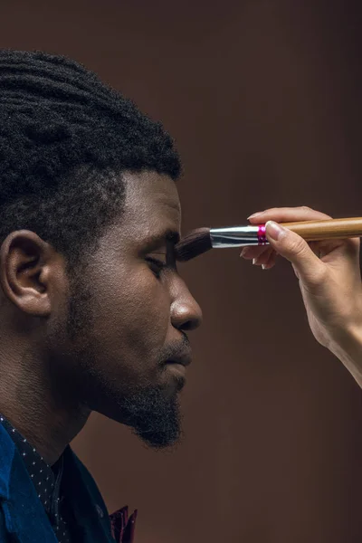 Imagem recortada de mulher aplicando pó de fundação com pincel de maquiagem no rosto de homem americano africano isolado em marrom — Fotografia de Stock Grátis