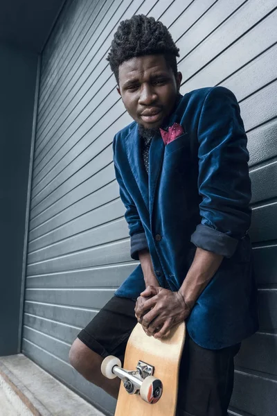 Hombre afroamericano con estilo en chaqueta azul apoyado en el monopatín y mirando a la cámara — Foto de Stock