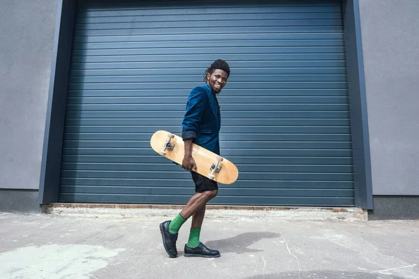 Stylish african american man in blue jacket with skateboard walking on street — Stock Photo, Image