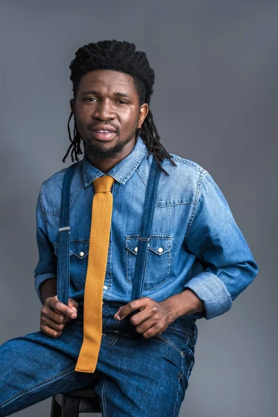 Handsome african american man sitting on chair and holding suspenders isolated on gray — Free Stock Photo