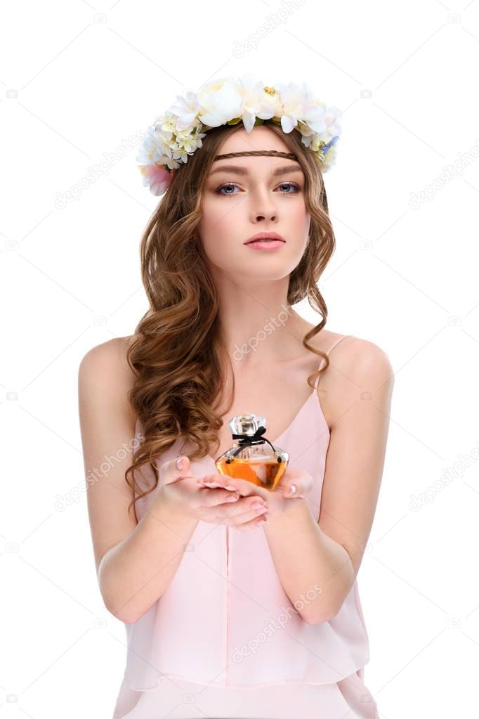 attractive young woman in floral wreath holding bottle of perfume isolated on white