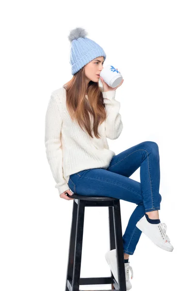 Belle femme en bonnet tricoté buvant du café assis sur tabouret, isolé sur blanc — Photo de stock