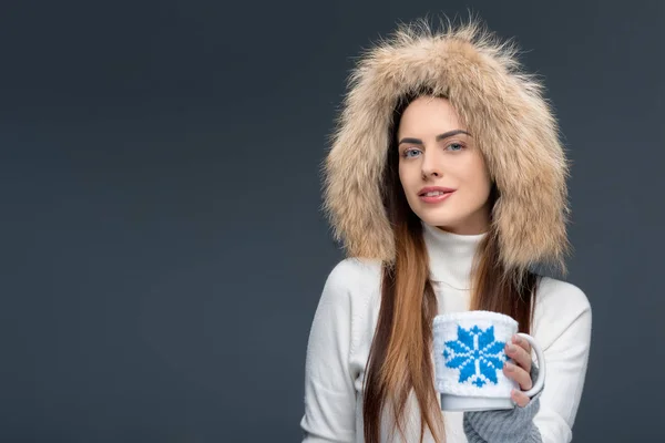 Belle femme en chapeau de fourrure et tenue d'hiver tenant une tasse de café, isolée sur gris — Photo de stock