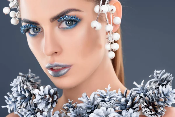 Retrato de modelo atractivo con accesorio para el cabello, conos de pino navideño, maquillaje de invierno y purpurina, aislado en gris - foto de stock