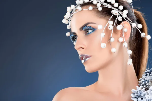 Modelo atractivo con accesorio para el cabello, conos de pino de Navidad, maquillaje de invierno y brillo, aislado en azul - foto de stock