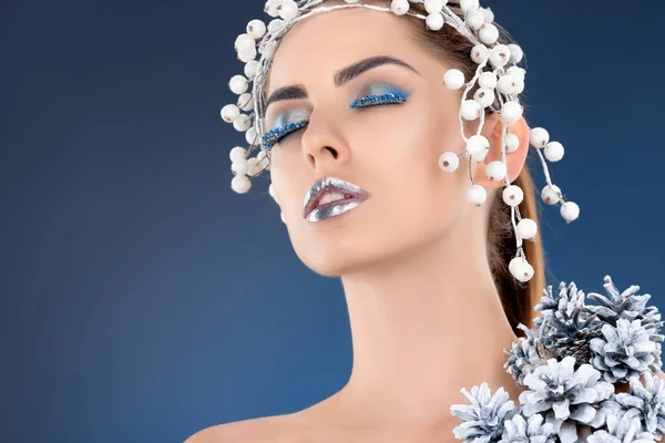 Portrait de beau modèle avec accessoire capillaire, cônes de pin de Noël, maquillage d'hiver et paillettes posant pour le tournage de mode, isolé sur bleu — Photo de stock