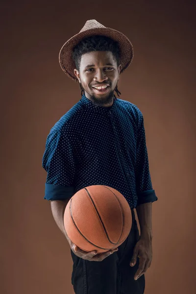 Американский спортсмен с улыбкой держит баскетбольный мяч изолированный на коричневом — стоковое фото