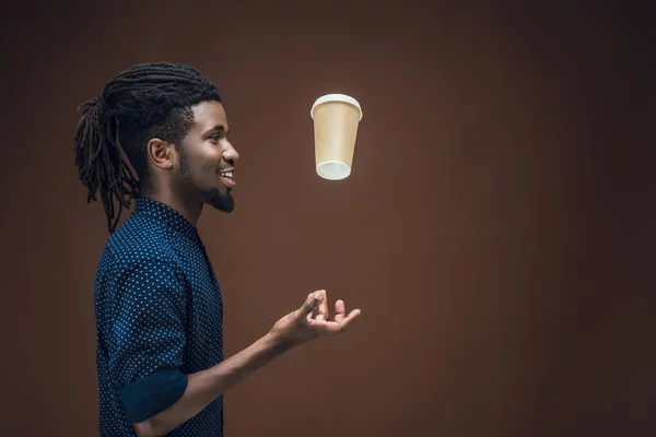 Side view of african american man tossing up coffee in paper cup isolated on brown — Stock Photo