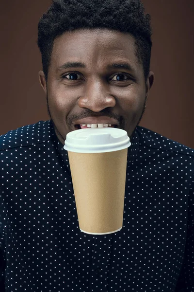 African american man holding disposable coffee cup with teeth isolated on brown — Stock Photo