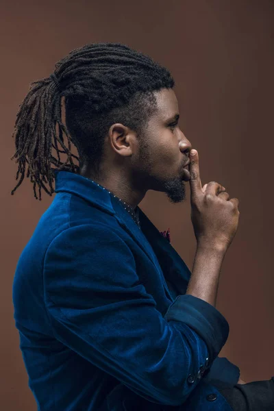 Боковой вид африканского мужчины, показывающего жест молчания, изолированный на коричневом фоне — стоковое фото
