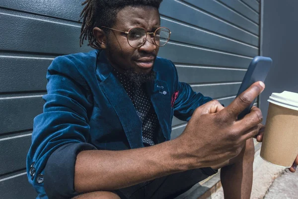 Élégant homme afro-américain veste bleue tenant café pour aller et en utilisant smartphone — Photo de stock