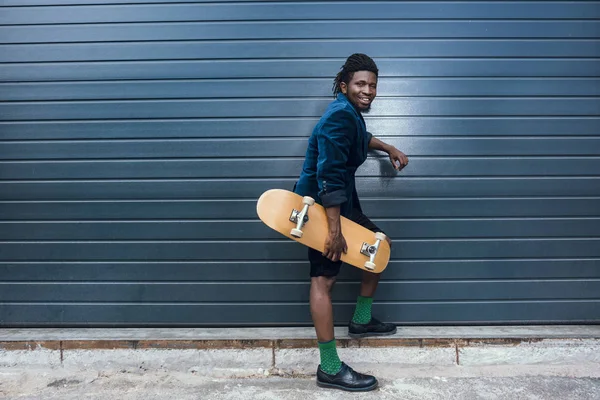 Stylish african american man in blue jacket holding skateboard and leaning on wall — Stock Photo