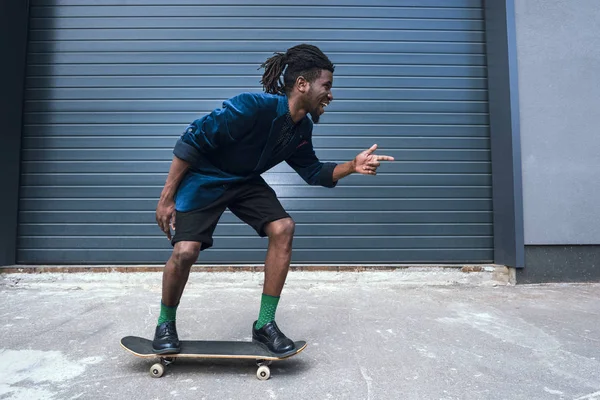 Élégant homme afro-américain en veste bleue patiner sur la rue et pointant sur quelque chose — Photo de stock