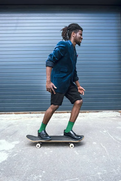 Vista lateral del hombre afroamericano con estilo en chaqueta azul patinaje en la calle - foto de stock