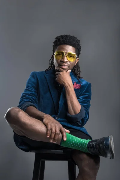 Hombre afroamericano con estilo en chaqueta azul y gafas de sol amarillas aisladas en gris - foto de stock