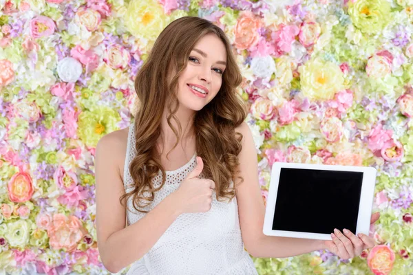 Hermosa mujer joven con la tableta que muestra el pulgar hacia arriba en el fondo floral - foto de stock