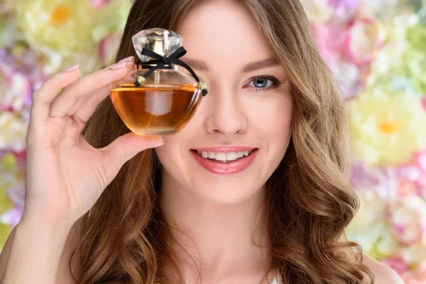 Retrato de cerca de una joven sonriente cubriendo un ojo con una botella de perfume - foto de stock