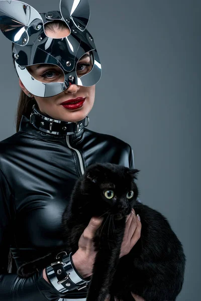 Mulher pervertida em traje sexy e máscara segurando gato preto isolado em cinza — Fotografia de Stock
