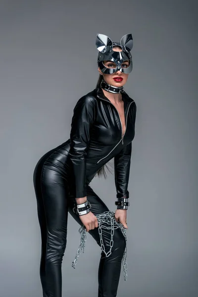 Mujer joven seductora con traje de gato y cadena de sujeción de máscara aislada en gris - foto de stock