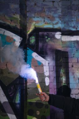 cropped view of hand with smoke bomb against wall with graffiti at night clipart