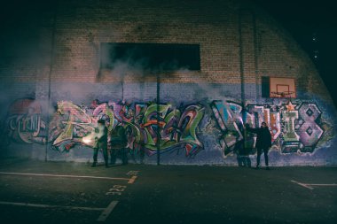 people holding smoke bombs and standing against wall with graffiti at night clipart