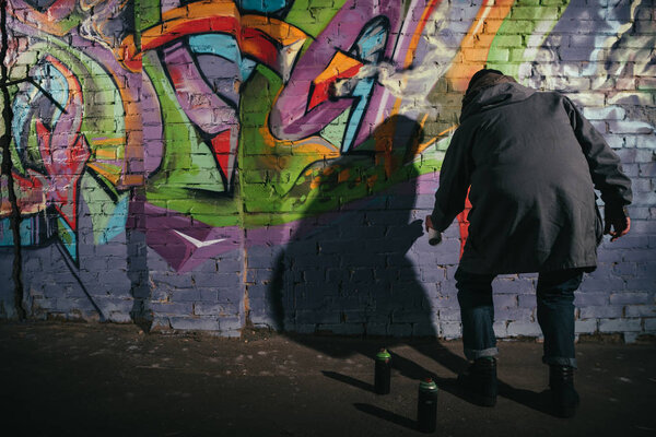 rear view of street artist painting graffiti with aerosol paint on wall at night