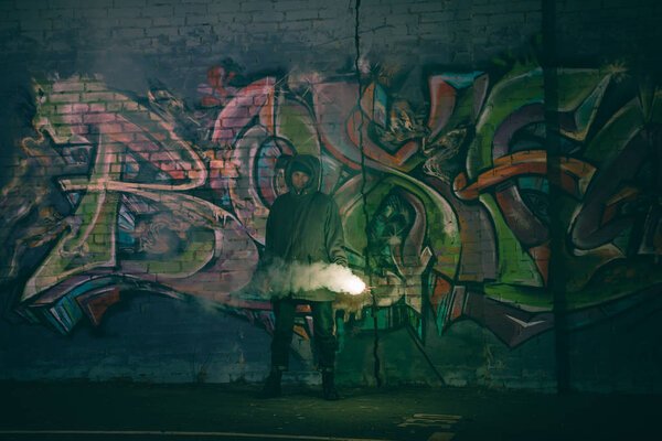 Человек, держащий дымовую бомбу и стоящий у стены с граффити ночью
