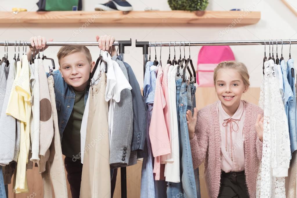 two smiling kids looking at camera while standing under cloth hanger at shop 