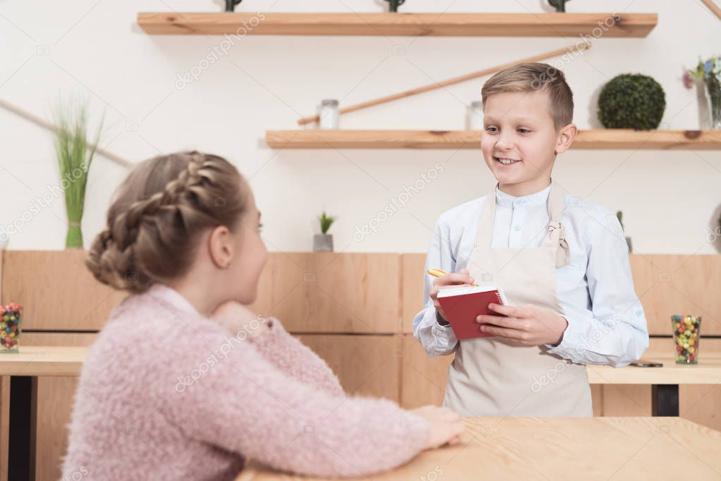 smiling little waiter making notes in notepad against child at cafe   