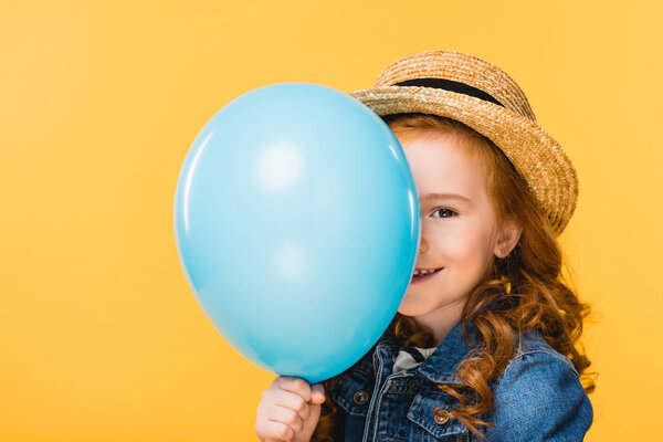 obscured view of smiling child covering face with balloon isolated on yellow