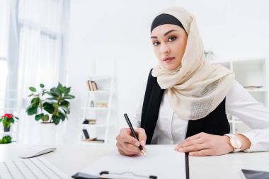 portrait of arabic businesswoman looking at camera while signing papers at workplace clipart