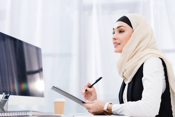 side view of smiling businesswoman in hijab with documents at workplace in office